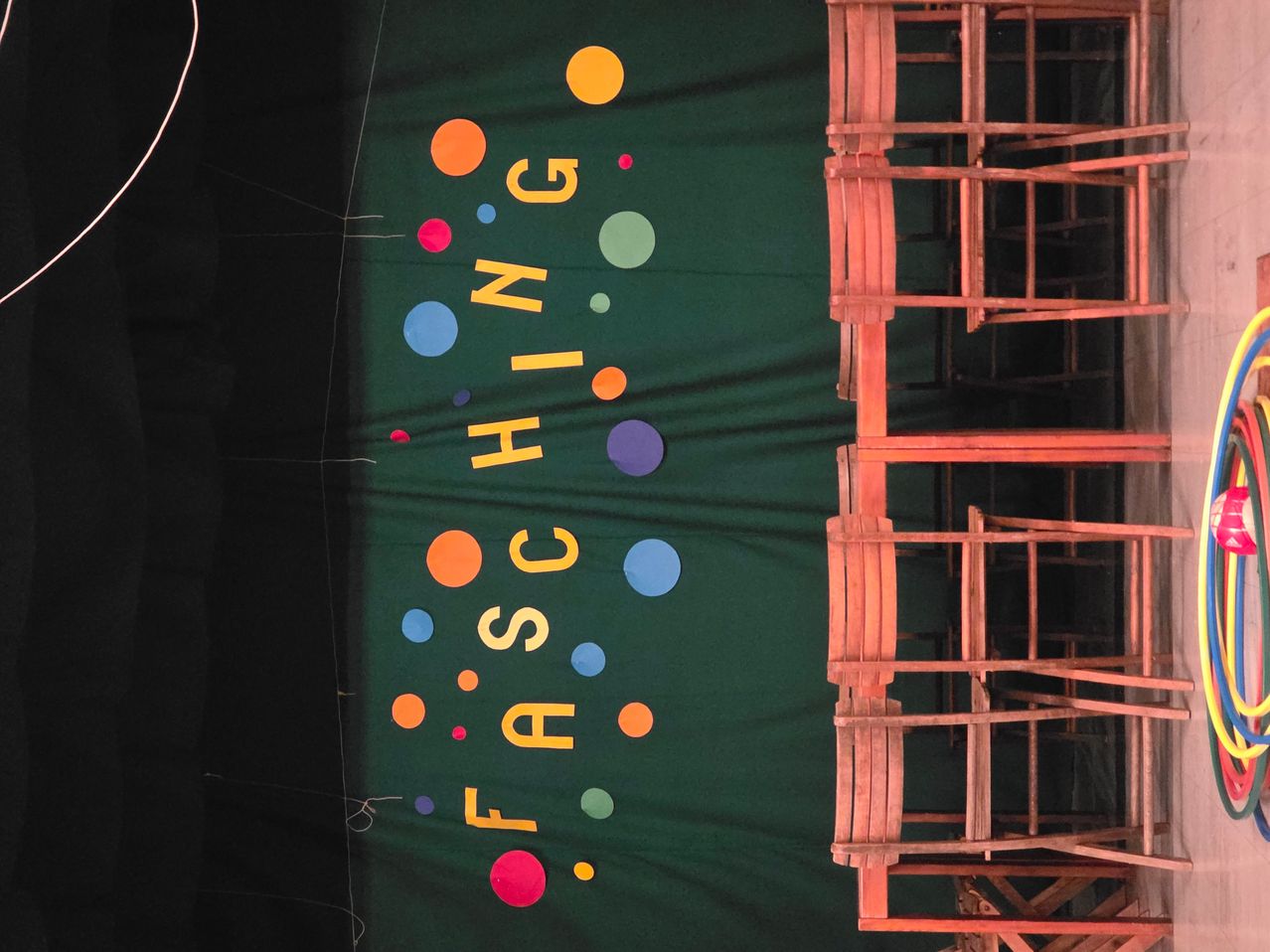 Ein grüner Hintergrund zeigt das Wort 'FASCING' in gelben Buchstaben, umgeben von mehrfarbigen Kreisen. Der Hintergrund ist leicht gefaltet, und eine rote Holzstruktur ist rechts sichtbar.