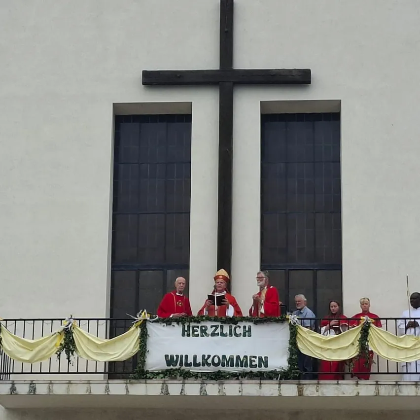 Eine Gruppe von Geistlichen in roten Roben steht auf einem Balkon mit einem Banner, das Herzlich Willkommen bedeutet. Ein großes Holzkreuz ist an einer weißen Wand hinter ihnen angebracht.