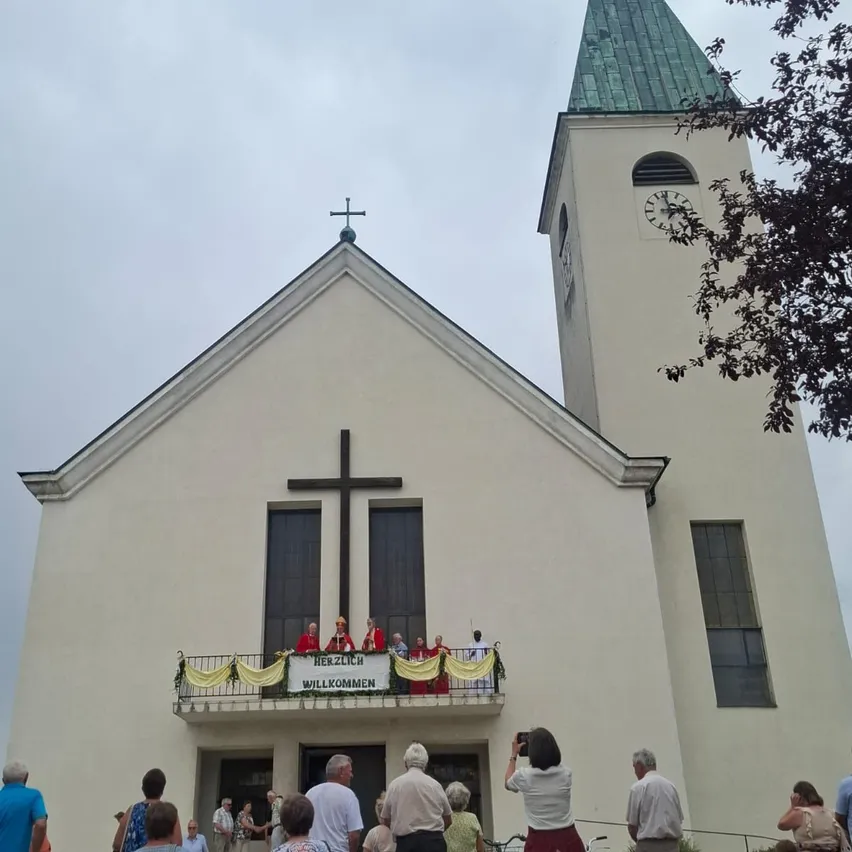 Eine Gruppe von Menschen versammelt sich vor einer Kirche, einige sind auf dem Balkon mit einem Banner, das 'Herzlich Willkommen' steht. Die Kirche hat ein Kreuz auf der Spitze und eine Uhr auf dem Turm.