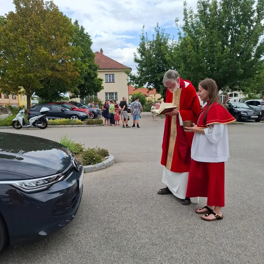 Ein Priester liest aus einem Buch, während ein junges Mädchen eine Schüssel in einem Parkplatz mit Menschen und Autos im Hintergrund hält.