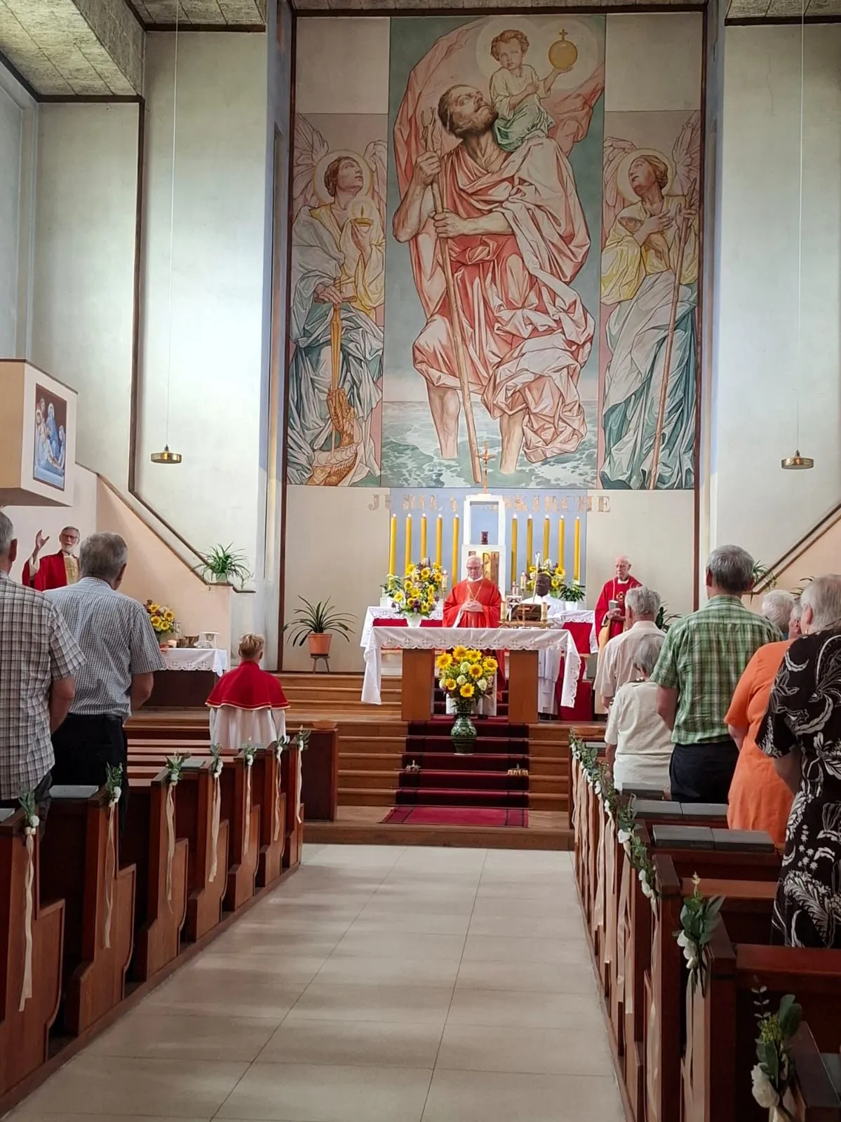 Ein Priester steht am Altar in einer Kirche, umgeben von Kerzen und Blumen, mit einem großen Wandbild hinter ihm. Menschen sind in den Kirchenbänken versammelt, einige tragen rote Gewänder.