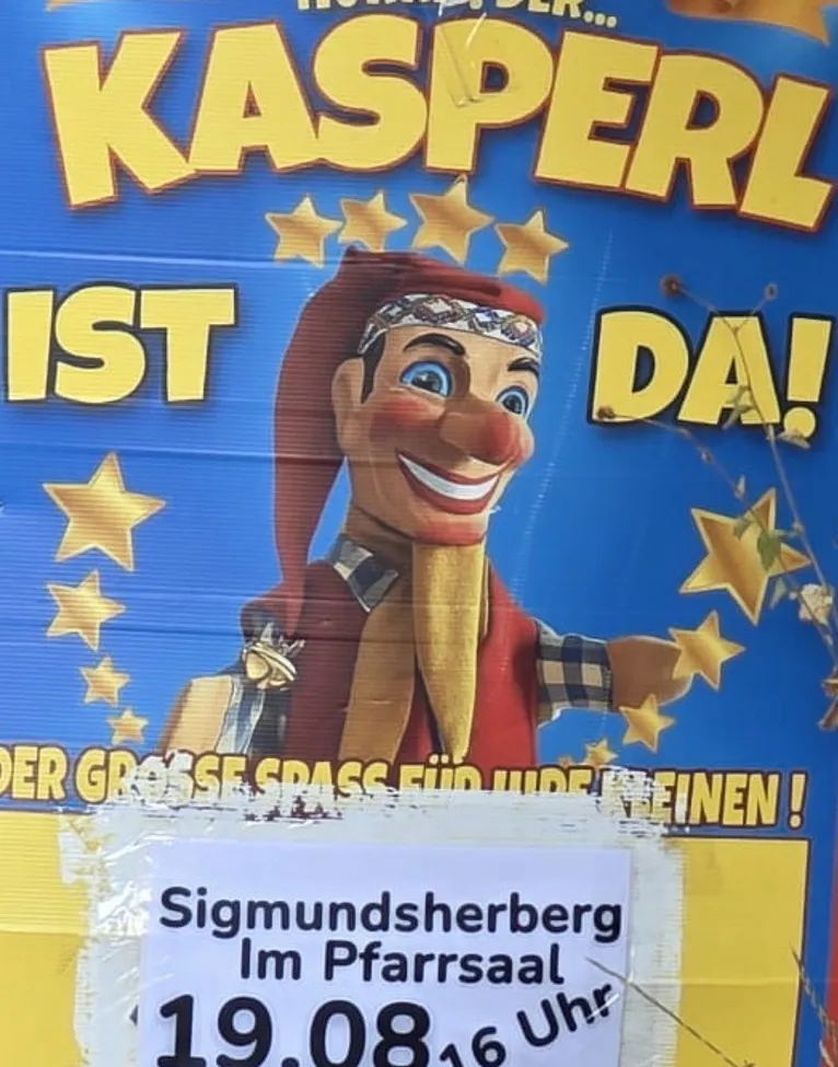 Ein Plakat für eine Puppenshow mit einer Figur namens Kasperl, die fröhlich lächelt und einen roten Hut trägt. Das Plakat hat einen blauen Hintergrund mit gelben Sternen und Text auf Deutsch.