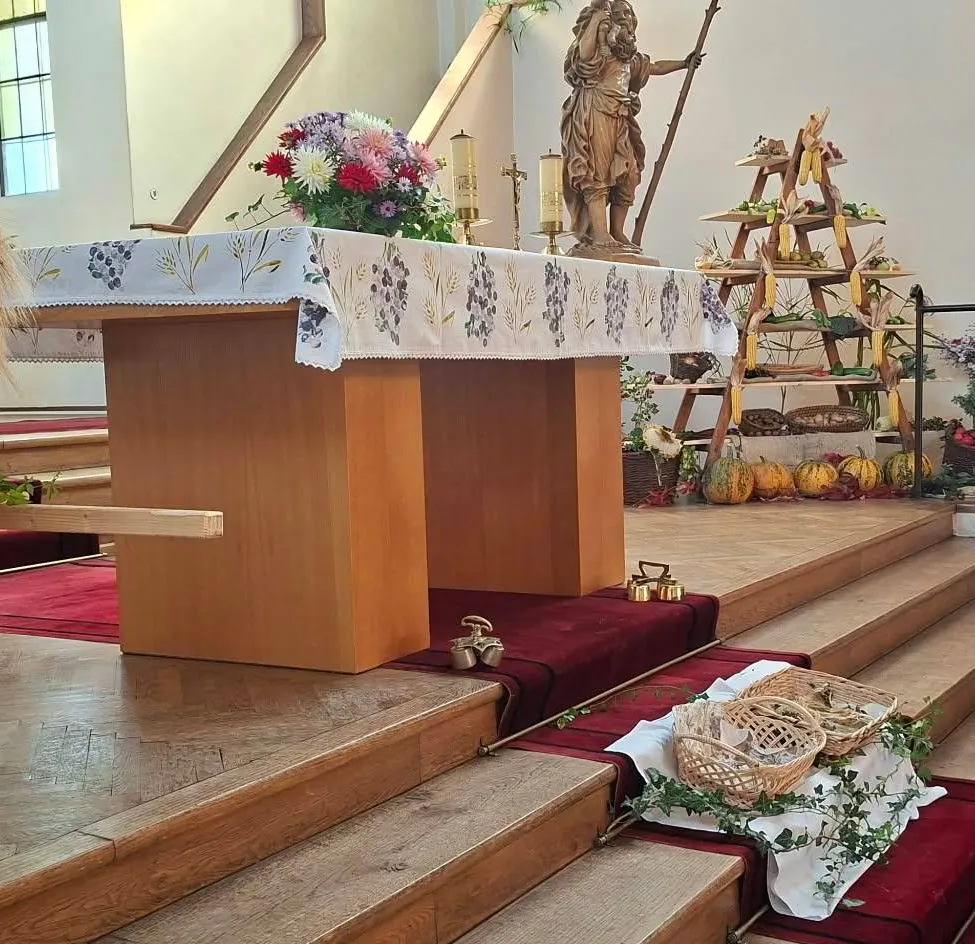 Ein hölzerner Altar mit einer weißen Tischdecke, Blumen und einer Statue einer Person mit einem Speer. Der Altar ist von Treppen und einem hölzernen Geländer umgeben.