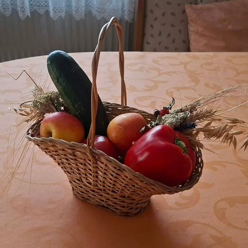 Ein Korb aus Weide mit frischem Obst und Gemüse, darunter Äpfel, Paprika und Gurken, steht auf einem Tisch mit gemusterter Tischdecke.