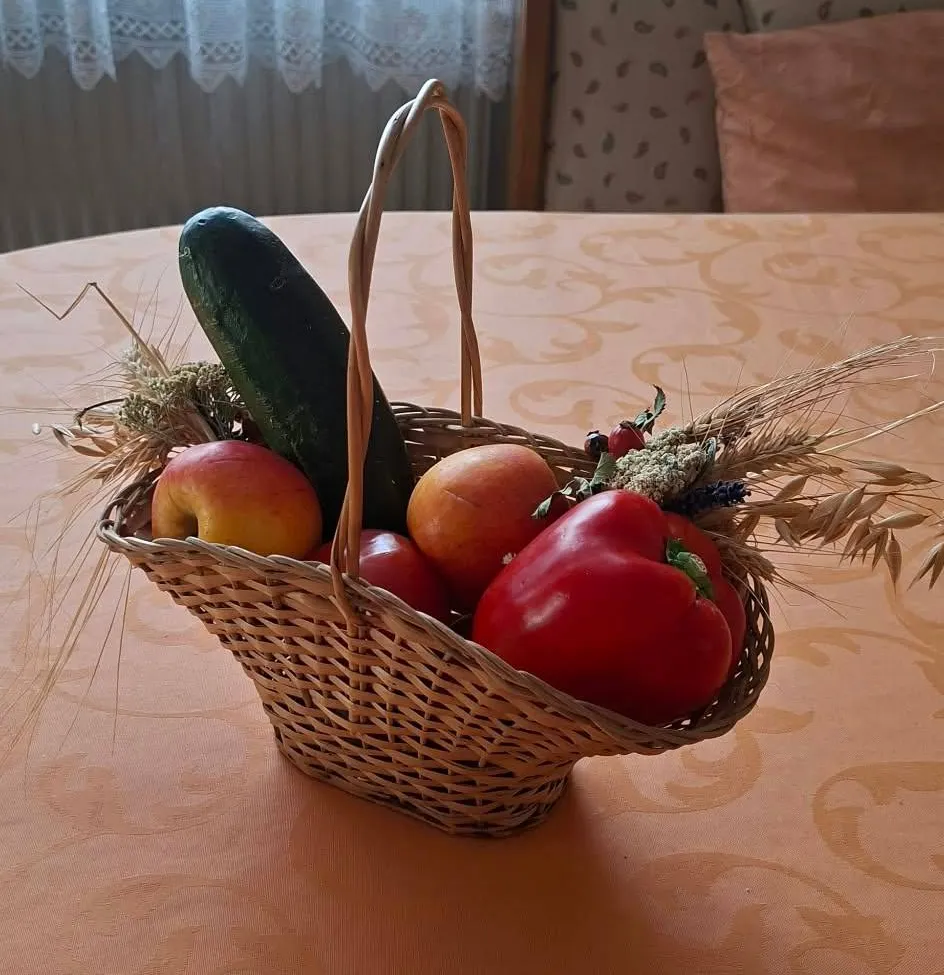 Ein Korb aus Weide mit frischem Obst und Gemüse, darunter Äpfel, Paprika und Gurken, steht auf einem Tisch mit gemusterter Tischdecke.