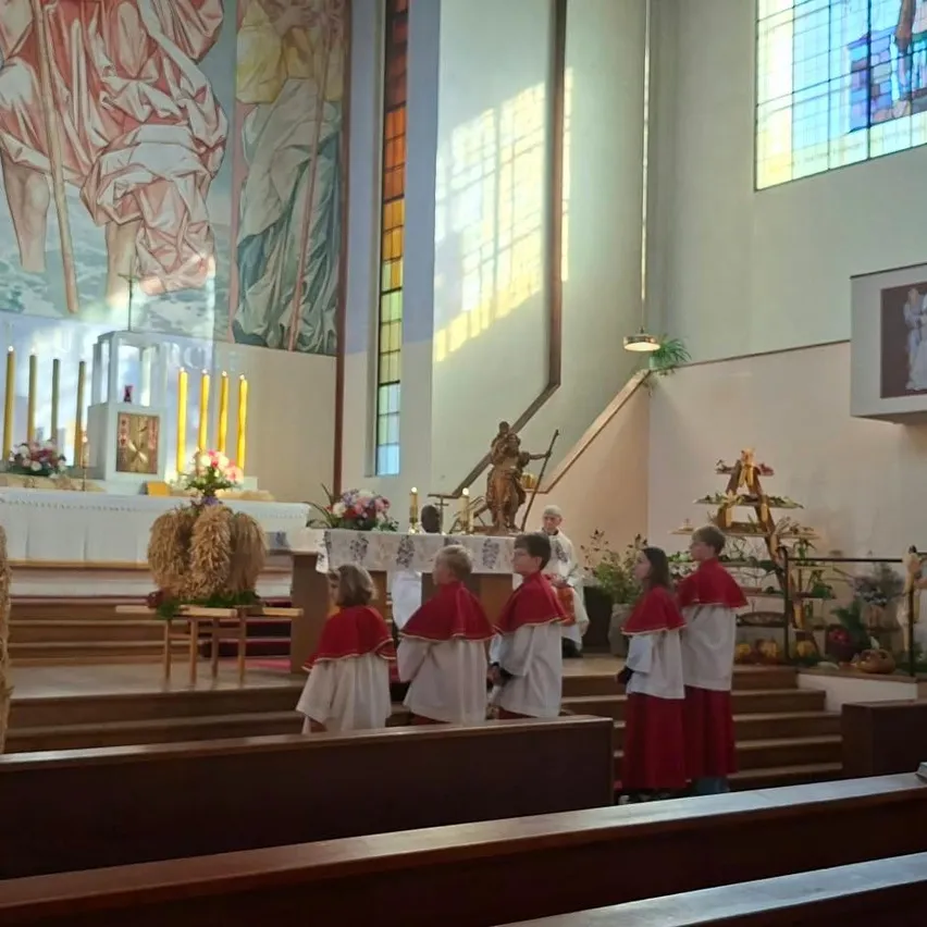 Ein Priester steht an einem Podium in einer Kirche und hält eine Predigt. Eine Gruppe von Menschen in roten und weißen Gewändern steht hinter ihm, und einige Leute sitzen auf den Kirchenbänken.