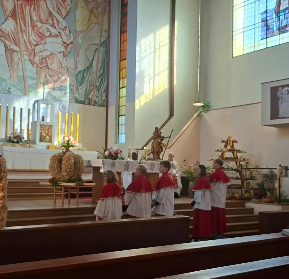Ein Priester steht an einem Podium in einer Kirche und hält eine Predigt. Eine Gruppe von Menschen in roten und weißen Gewändern steht hinter ihm, und einige Leute sitzen auf den Kirchenbänken.