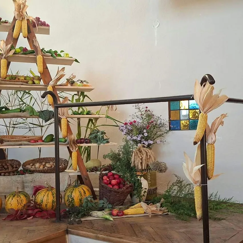 Eine holzerne Leiterstruktur, gefüllt mit verschiedenen Obst- und Gemüsesorten, darunter Kürbisse, Mais und Blumen, wird auf einem Holzboden ausgestellt.