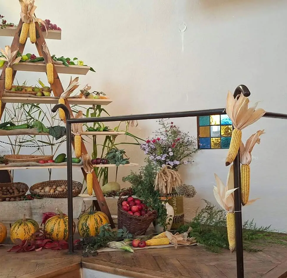 Eine holzerne Leiterstruktur, gefüllt mit verschiedenen Obst- und Gemüsesorten, darunter Kürbisse, Mais und Blumen, wird auf einem Holzboden ausgestellt.