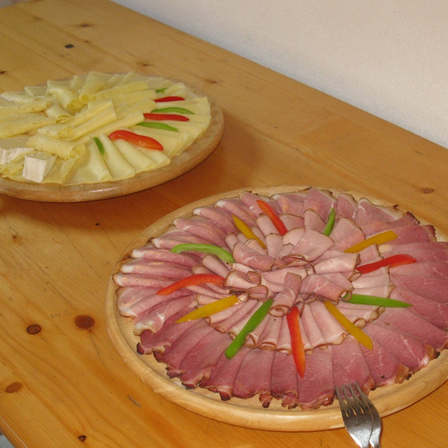 Zwei Holzplatten auf einem Holztisch mit verschiedenen Speisen. Eine hat Käse und Paprika, die andere hat Schinken und Paprika. Eine Gabel ist auf der Platte mit Schinken.