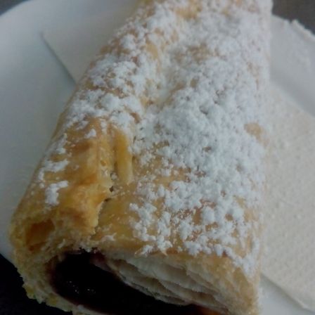 Ein Gebäck-Rolle mit blättrigen Schichten, bestreut mit weißem Pulver, wird auf einem weißen Teller präsentiert. Die Rolle ist aufgeschnitten und zeigt eine dunkle Füllung im Inneren.