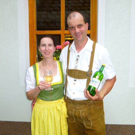 Ein Mann und eine Frau stehen draußen und halten Getränke. Der Mann ist in traditioneller deutscher Kleidung. Die Frau hält ein Glas Weißwein.