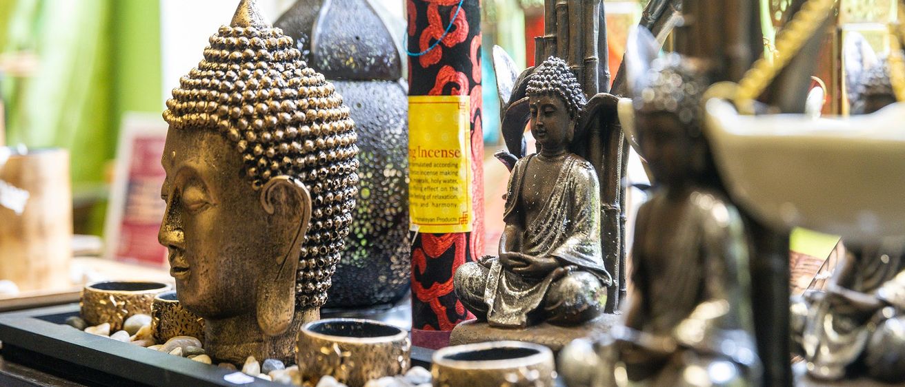Eine Sammlung von Buddha-Statuen und Räucherstäbchen, die auf einem Tisch ausgestellt sind. Die Statuen variieren in Größe und Pose, wobei eine in Meditationshaltung sitzt. Ein zylindrischer Räucherstab mit Etikett ist prominent ausgestellt.