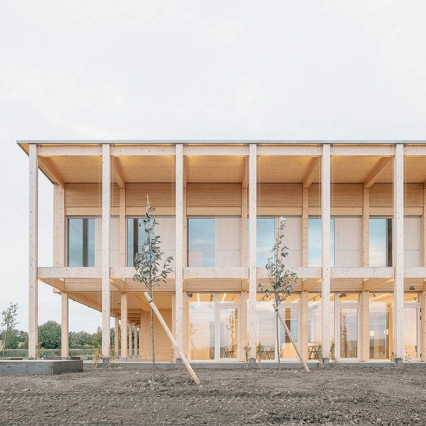 Ein modernes Gebäude mit Holzkonstruktion, Glasfenstern und großen Säulen. Vor dem Gebäude sind Bäume gepflanzt. Das Gebäude ist von einem Erdfeld umgeben.