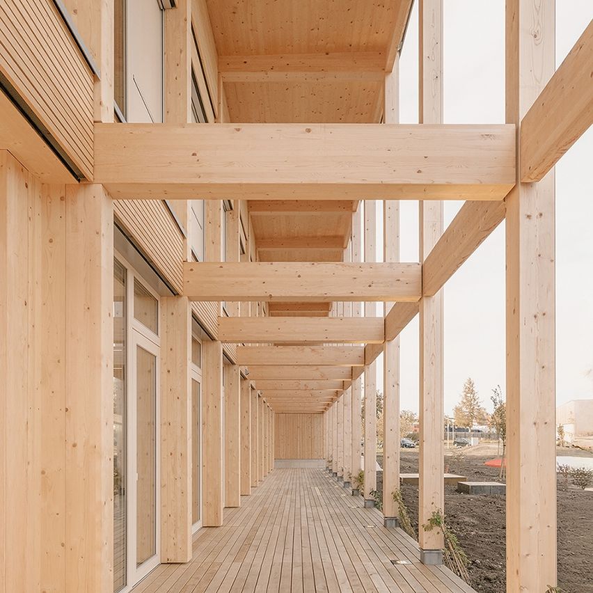 Ein leerer Holzkorridor mit Glastüren, Holzbalken und einem Holzboden. Die Wände sind aus Holz und es gibt kleine Pflanzen im Bereich. In der Ferne sind Bäume und ein Gebäude.