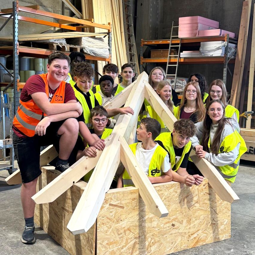 Eine Gruppe von Schülern in Warnwesten baut eine Struktur aus Holzbalken in einem Lagerhaus.