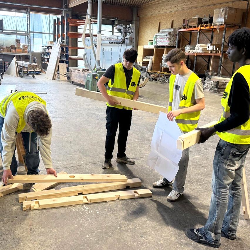 Vier Männer in Sicherheitswesten arbeiten in einem Lagerhaus an Holzbrettern. Einer hält ein Brett, ein anderer Papier, und zwei andere sind gebeugt. Lagerboxen stehen hinter ihnen auf Regalen.