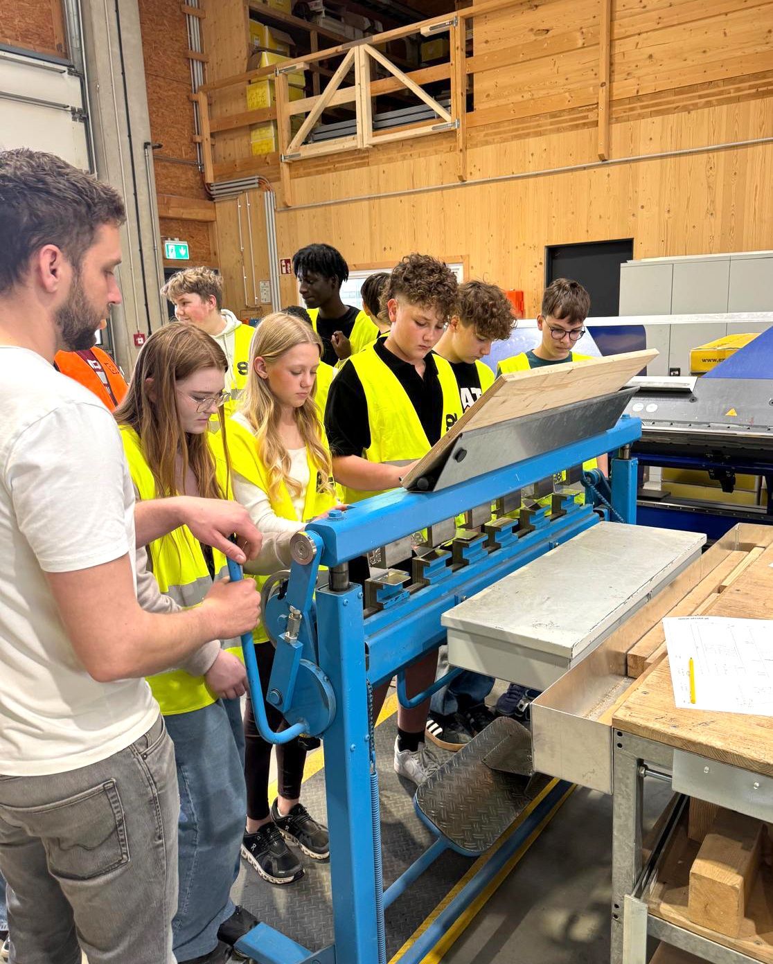 Eine Gruppe von Schülern in gelben Westen lernt in einer Werkstatt, eine Maschine zu bedienen. Ein Mann leitet sie, während sie einen blauen Hebel drücken.