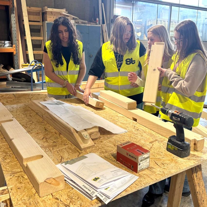 Vier Frauen in gelben Sicherheitswesten arbeiten an Holzbrettern auf einem Holztisch, eine hält eine Bohrmaschine.