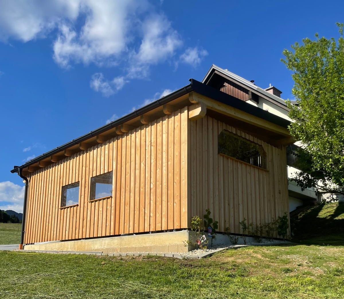 Ein modernes Holzhaus mit drei Fenstern, umgeben von saftigem Gras und einem blauen Himmel mit Wolken.
