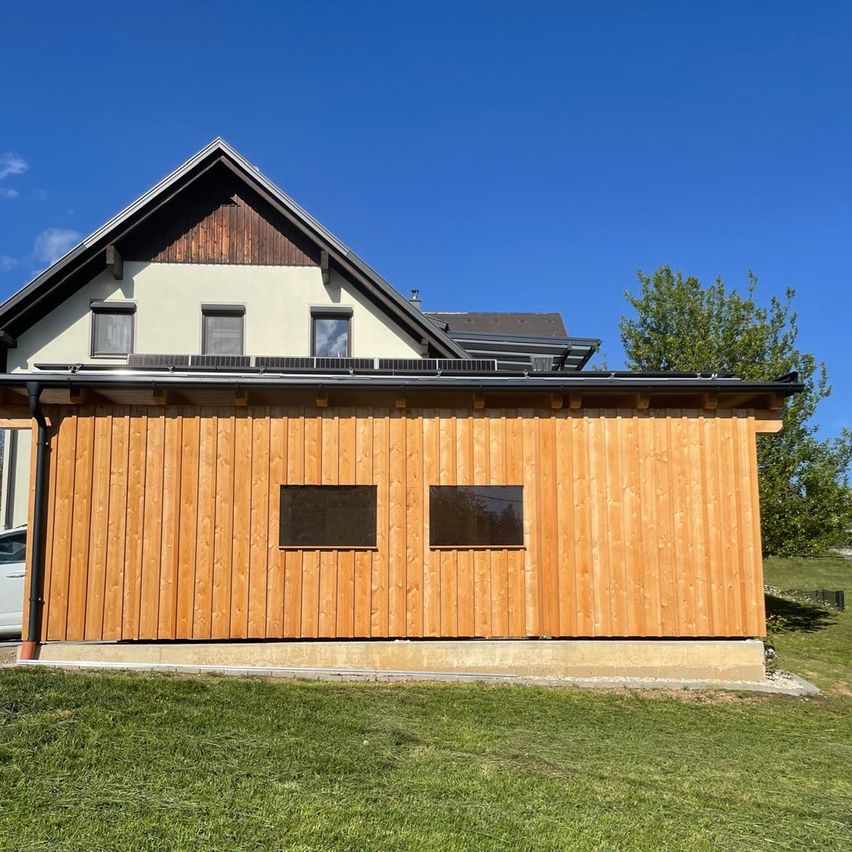 Ein modernes Haus mit hölzerne Außenwand und Solarpanels auf dem Dach, umgeben von einem Rasen und einem Baum.