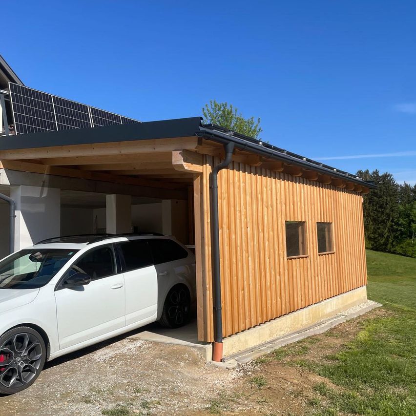 Ein weißer SUV ist unter einem hölzernen Carport mit Solarmodulen auf dem Dach geparkt. Der Carport ist an ein Haus angeschlossen.