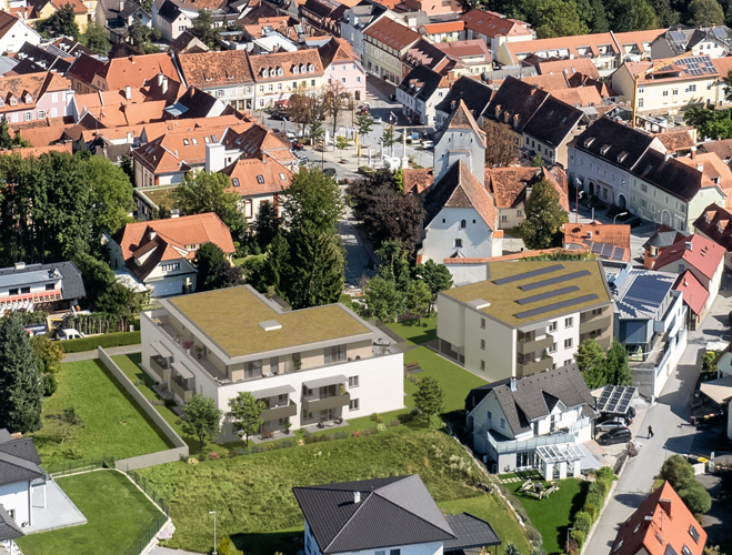 Aerial view of a residential area with various houses, some featuring solar panels, surrounded by trees and green grass, with a central road and a building under construction.