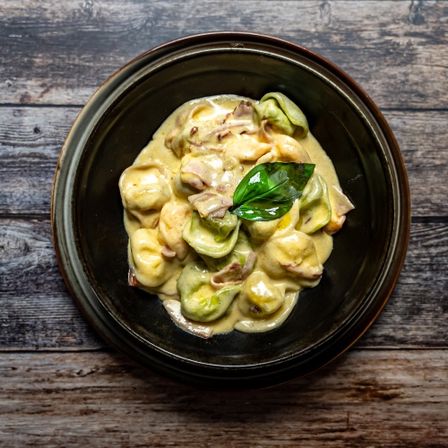 Eine Schüssel mit Tortellini-Pasta mit cremiger Sauce und mit Basilikumblatt garniert, auf einem Holztisch.