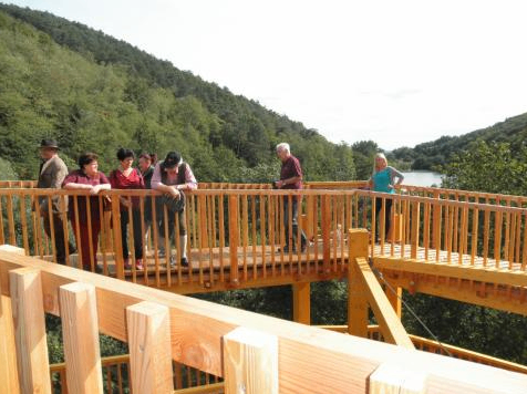 Sechs Personen stehen auf einer Holzbrücke über einem See, mit üppigen grünen Bergen im Hintergrund.