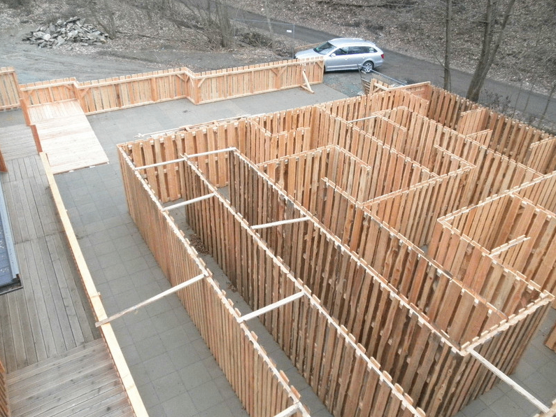 Luftaufnahme einer Holzlabyrinth-Struktur im Aufbau, mit einem silbernen Auto, das in der Nähe auf einer asphaltierten Straße geparkt ist.