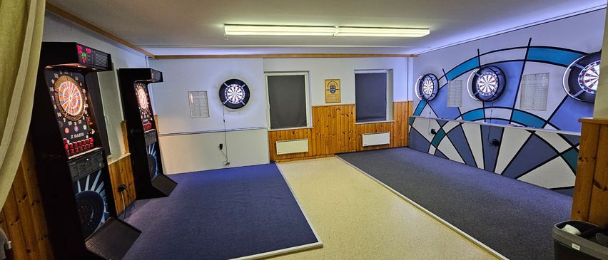 Ein leerer Raum mit Holzwänden, zwei Dartscheiben, zwei Fenstern und einer weißen Wand. Eine Dartscheibe ist an der Wand montiert, während die andere an einer blauen Wand montiert ist.