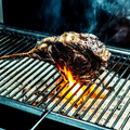 Ein Stück Fleisch, möglicherweise ein Lammkarree, wird auf einem Grill gegrillt. Das Fleisch steht in Flammen und hat eine goldbraune Farbe. Es gibt kleine Steine und Kohlen auf dem Grill. Der Rauch kommt von der Kohle.