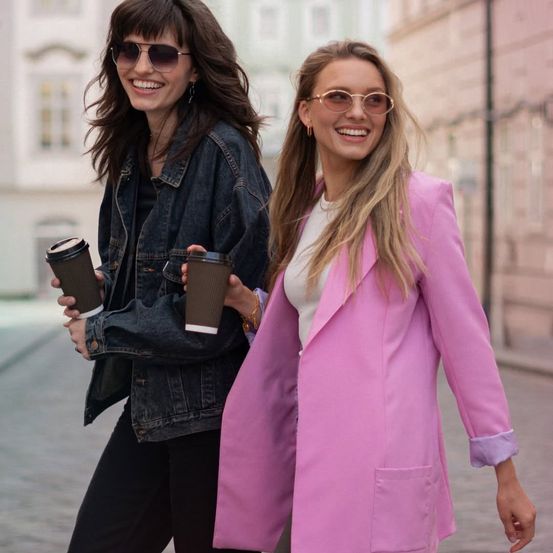 Zwei Frauen gehen eine Stadtstraße entlang, eine trägt eine Jeansjacke und die andere einen rosa Blazer, beide halten Kaffeebecher und lächeln.