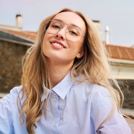 Eine Frau mit blonden Haaren lächelt und trägt eine Brille. Sie ist in einem hellblauen Hemd gekleidet und steht draußen.