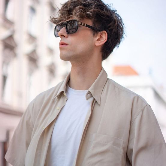 Ein Mann mit lockigem Haar, der ein weißes T-Shirt und eine beige Jacke trägt, steht auf einer Stadtstraße und schaut nach oben.