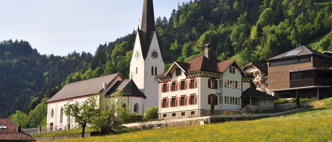 Eine malerische Kirche mit einem Turm und einem Uhrenturm steht in einem Dorf, umgeben von einem Feld gelber Blumen und einem bewaldeten Hang.