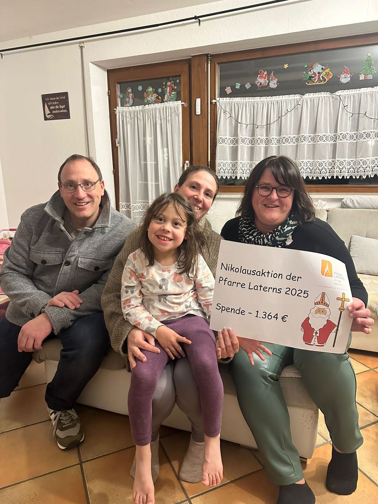 Drei Erwachsene und ein junges Mädchen sitzen auf einer Couch und halten ein Schild mit einer Zeichnung des Heiligen Nikolaus. Auf dem Schild steht 'Nikolausaktion der Pfarre Laterns 2025 Spende - 1.364 €'. Dahinter befindet sich ein Fenster mit weißen Vorhängen, das mit Weihnachtsdekorationen geschmückt ist.
