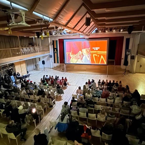 Ein großer Innenraum mit Bühne, großer Leinwand und vielen Menschen in Stühlen. Die Leinwand zeigt einen Zeichentrickcharakter.