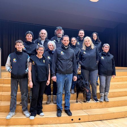 Eine Gruppe von Menschen, die schwarze Jacken tragen, posiert für ein Foto auf hölzernen Stufen mit einem Hund vor ihnen.