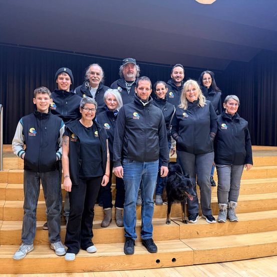 Eine Gruppe von Menschen, die schwarze Jacken tragen, posiert für ein Foto auf hölzernen Stufen mit einem Hund vor ihnen.