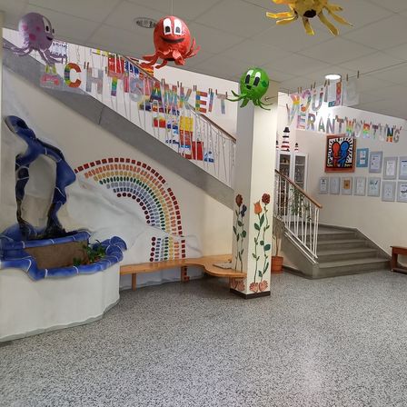 Ein dekorierter Innenraum mit einer Treppe, einer Regenbogenwand und verschiedenen hängenden Dekorationen. Eine kleine Bank und Topfpflanzen stehen auf dem Boden. Die Decke hat hängende Dekorationen und Lichter.