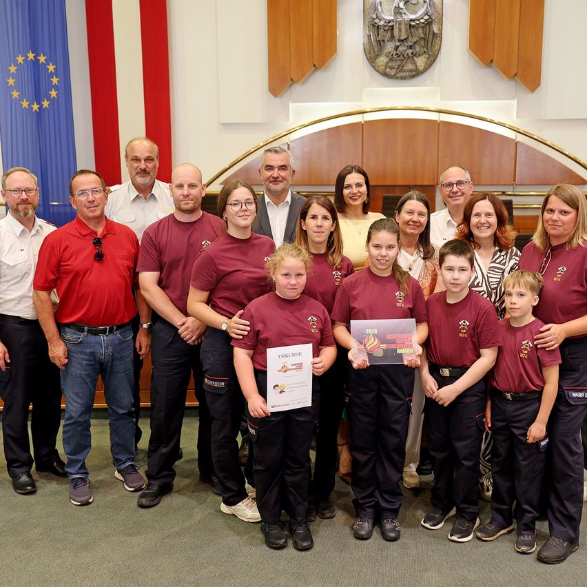 Eine Gruppe von Menschen in roten Uniformen steht in einem Raum, einige halten Zertifikate. Hinter ihnen stehen Erwachsene mit einer Flagge und einer dekorativen Wand.