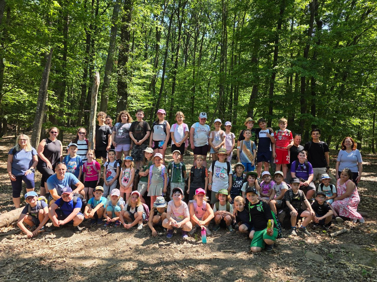 Eine Gruppe von Menschen, einschließlich Erwachsener und Kinder, posiert für ein Foto in einem bewaldeten Gebiet. Sie sind lässig gekleidet, einige mit Hüten und Sonnenbrillen. Bäume umgeben sie und der Boden ist mit Schmutz bedeckt.