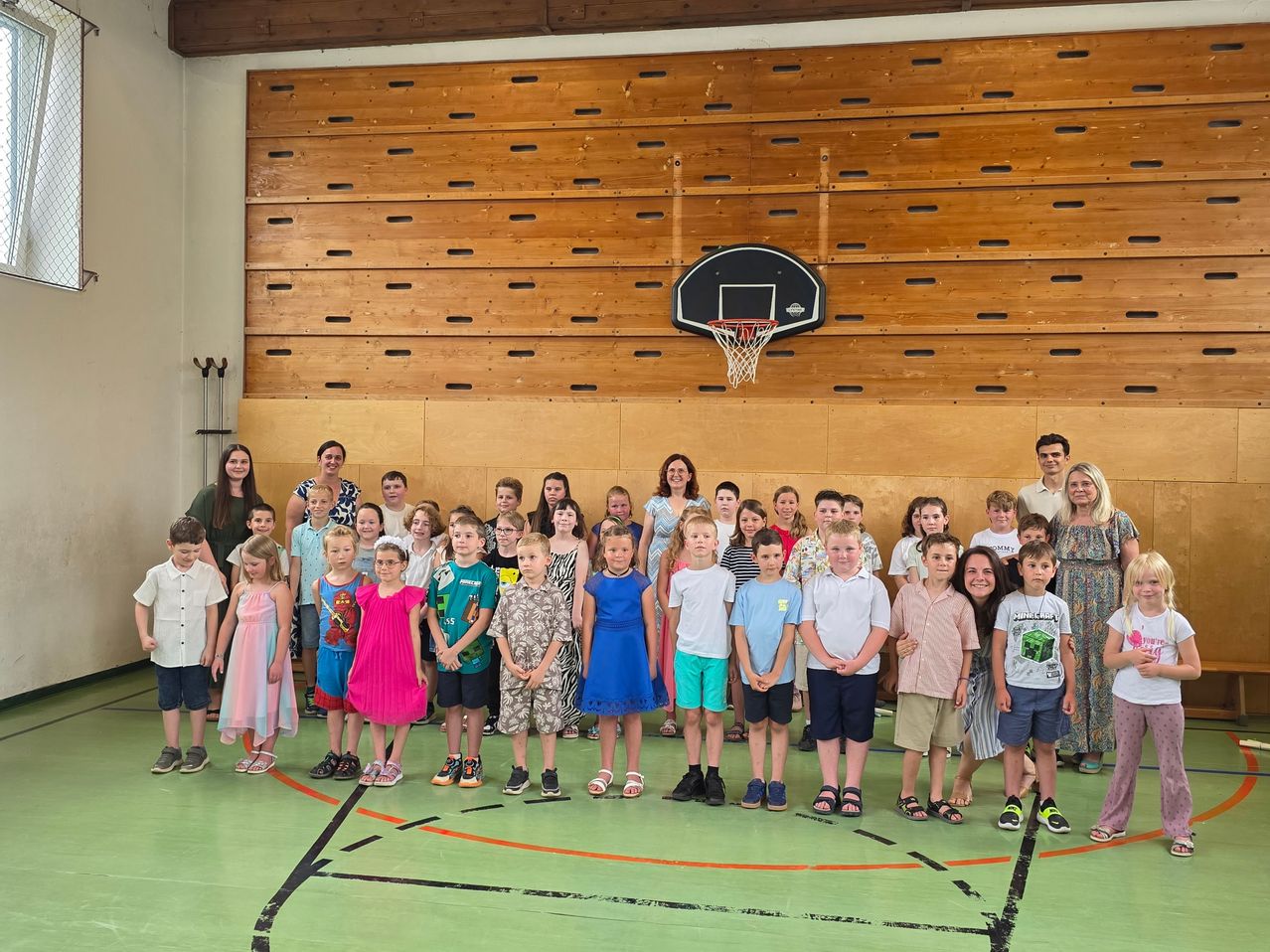 Eine Gruppe von Kindern und Erwachsenen posiert für ein Foto in einer Turnhalle, mit einem Basketballkorb im Hintergrund.