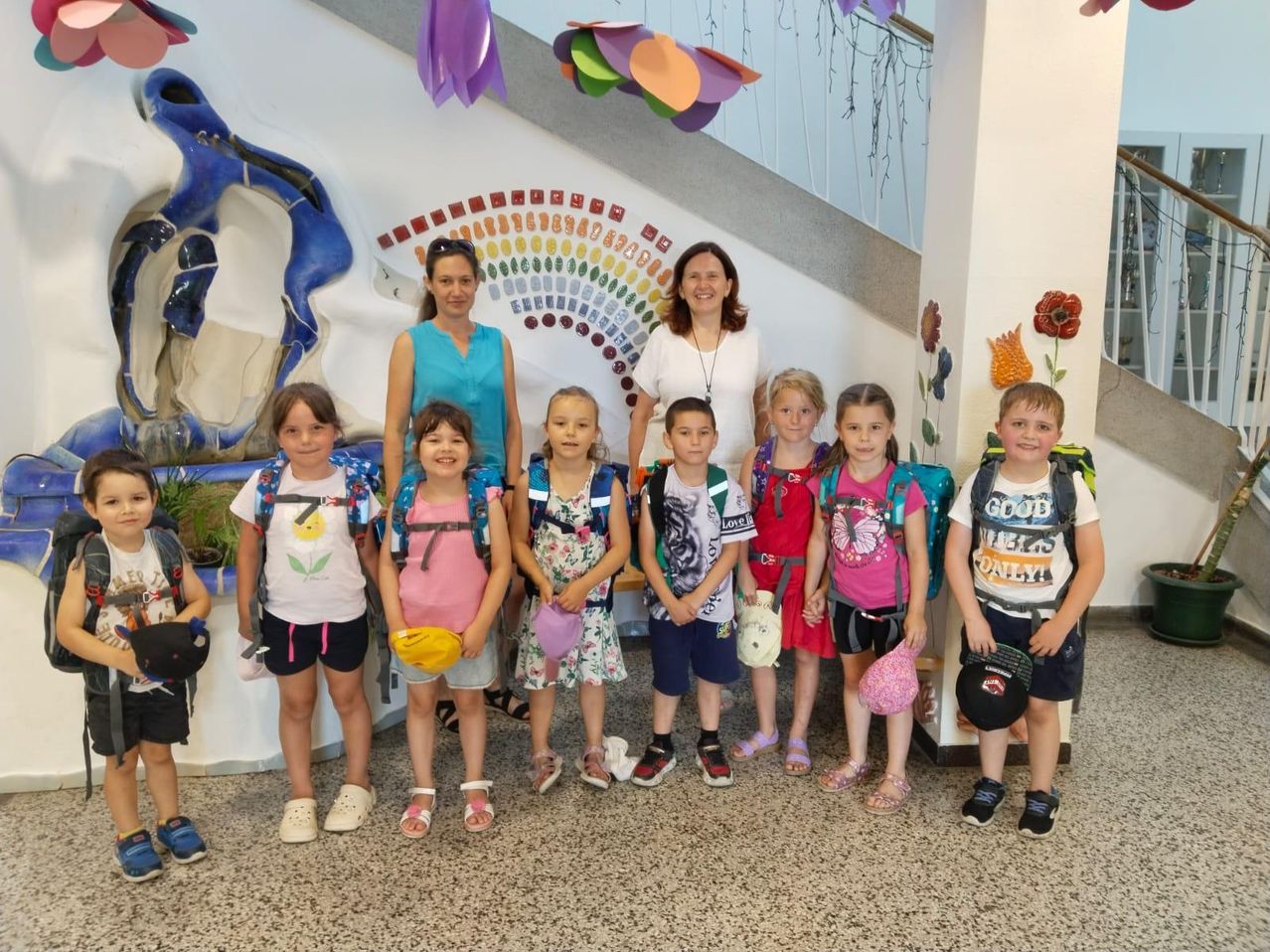 Eine Gruppe von Kindern mit ihren Lehrern steht vor einer bunten Wand und einer Treppe. Sie tragen Rucksäcke und halten Hüte und lächeln für ein Foto.