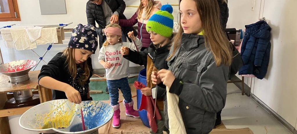 Eine Gruppe von Kindern und Erwachsenen versammelt sich um einen Topf mit Farbe. Sie mischen Farbe und schauen sich gegenseitig an.