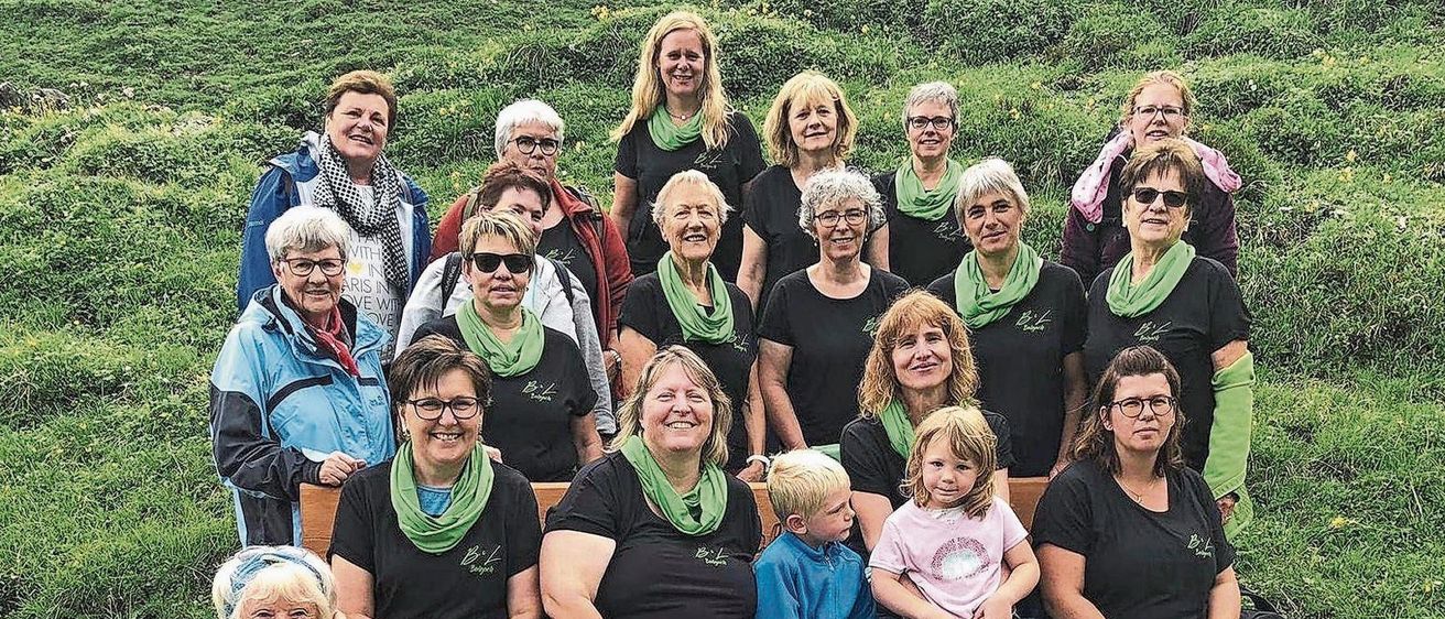 A group of women in black t-shirts and green scarves poses for a photo in a grassy field with a wooden bench and a child sitting.