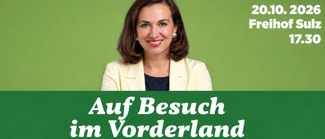 Eine Frau steht vor einem grünen Hintergrund mit den Worten 'Auf Besuch im Vorderland Alma Zadic' in weißer Schrift. Sie lächelt und trägt einen hellen Blazer und schwarze Hosen.