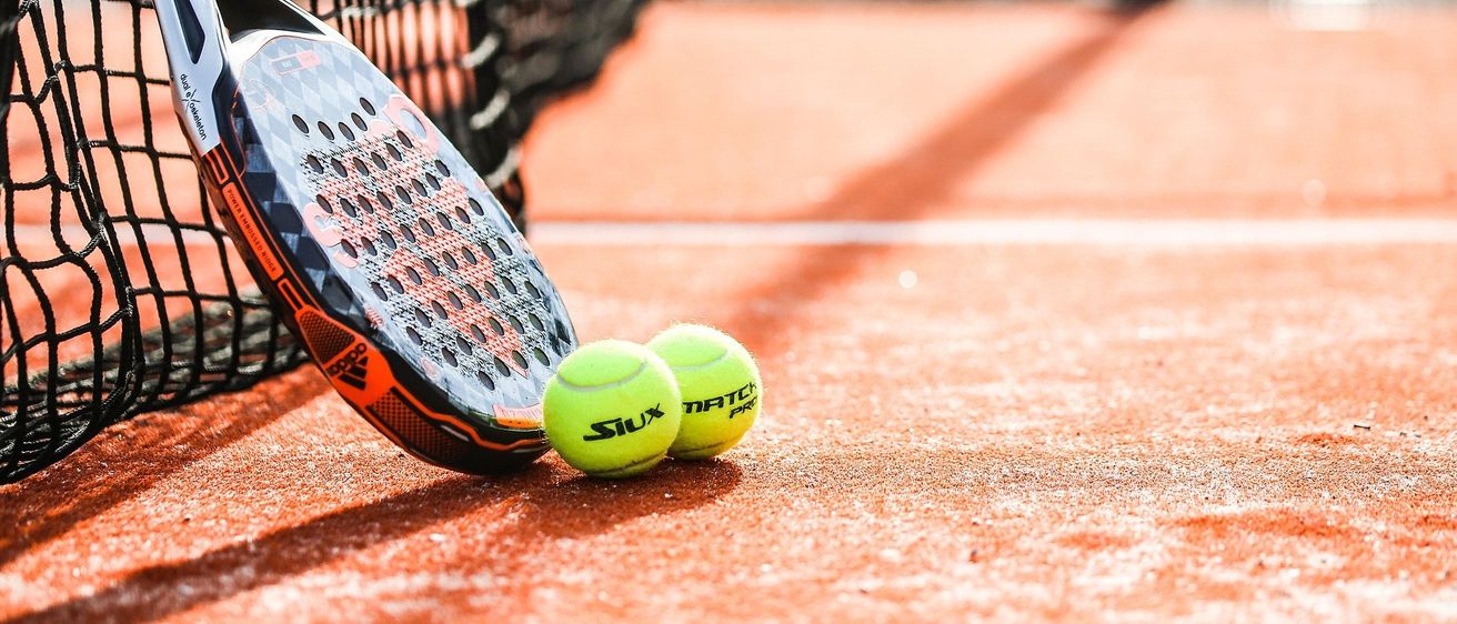 Ein Tennisschläger liegt neben zwei Tennisbällen auf einem Tennisplatz.