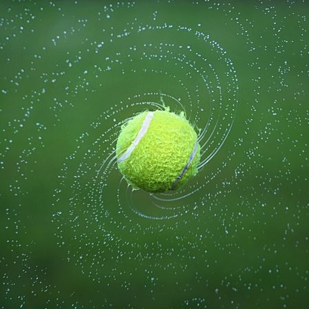 Ein Tennisball ist in der Luft, umgeben von Wassertropfen. Der Ball hat eine weiße Linie und einen lila Streifen. Der Hintergrund ist grün.